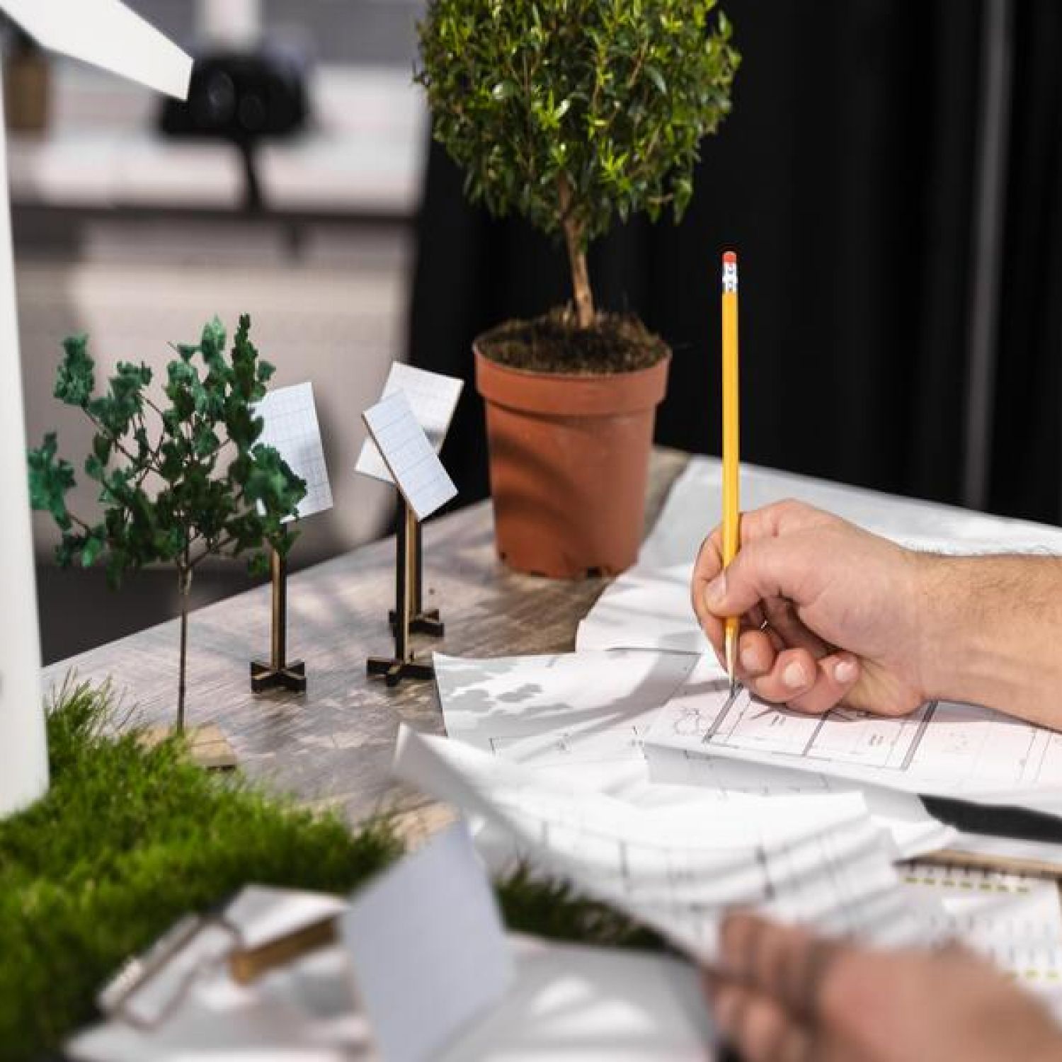 Mão de um homem branco escrevendo em um documento sob uma mesa de madeira ao lado de miniaturas de plantas de plástico