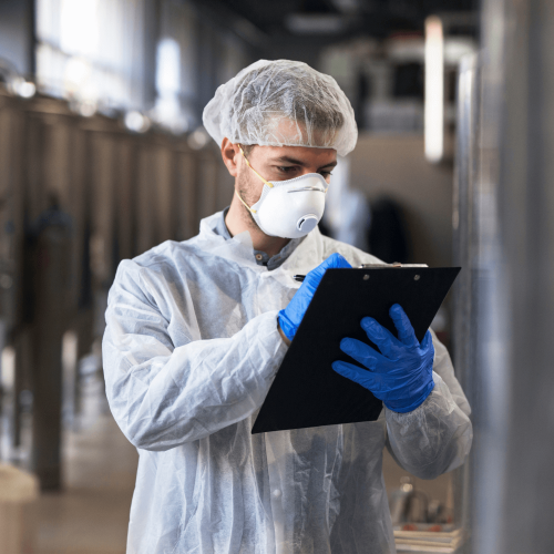 Homem de jaleco, touca e máscara checando produtos químicos em uma indústria